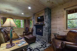 Interior acogedor de una cabaña con chimenea de piedra, mobiliario de cuero y un televisor montado en la pared.