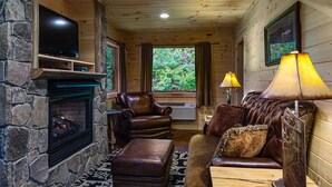 A cozy cabin interior with a stone fireplace, leather furniture, and a large window with a view of greenery. - Black Bear Pond log cabin (Almond)