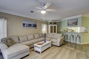 A living room with a beige sofa, a white coffee table, and a ceiling fan. There is a kitchen area with a bar and stools. - Sunset Rentals | Ocean Breeze 18 | Dog Friendly (Hilton Head Island)