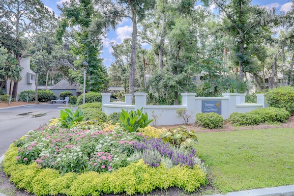 A landscaped garden with a variety of plants and flowers, a white building with a sign, and a street in the background. - Sunset Rentals | Ocean Breeze 18 | Dog Friendly (Hilton Head Island)