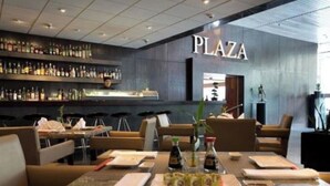 Een moderne bar met een 'PLAZA' bord, zitgelegenheden en diverse flessen op planken.