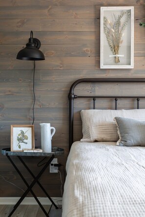 Un dormitorio con pared de madera, estructura de cama metálica, mesita de noche con lámpara, impresión botánica enmarco y jarra blanca.