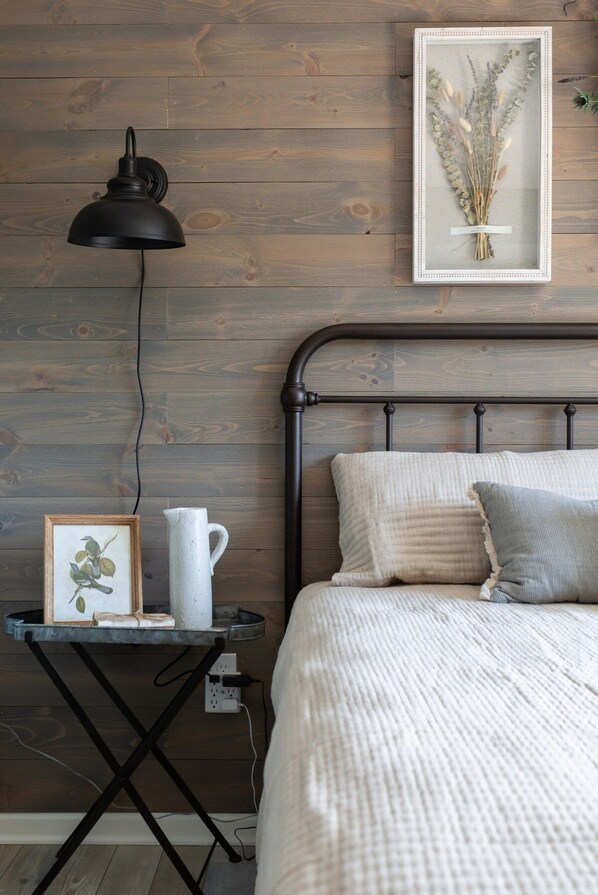 Un dormitorio con pared de madera, estructura de cama metálica, mesita de noche con lámpara, impresión botánica enmarco y jarra blanca.