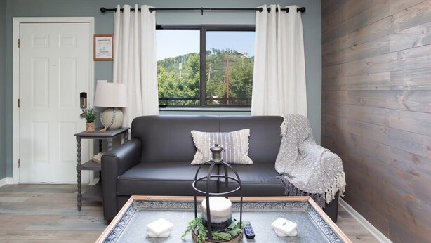 Una sala de estar con un sofá de cuero negro, una mesa de centro con velas y un adorno, una ventana con cortinas y una pared de madera.