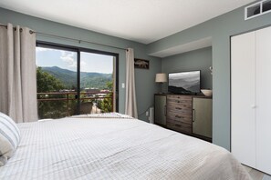 Un dormitorio con una cama grande, un aparador, un televisor y vistas a montañas desde la puerta del balcón.