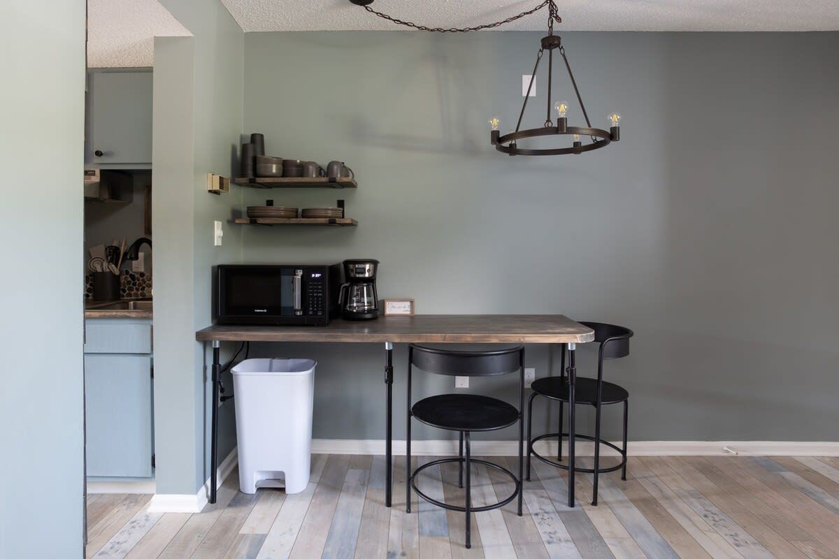 Une cuisine avec une table en bois, deux chaises noires, un luminaire suspendu et une poubelle.