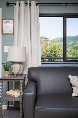 Una habitación con un sofá de cuero negro, una lámpara, un plano de evacuación enmarcado y vistas a un paisaje verde a través de una ventana.