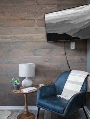 Una habitación con pared de madera, televisor montado, sillón de cuero azul, mesita de madera con una lámpara y una planta en maceta.