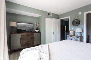 A bedroom with a bed, a dresser, a television, and a door leading to a kitchen.