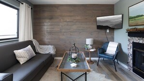 Una sala moderna con un sofá de cuero oscuro, una mesa de centro con una vela, un sillón azul, una chimenea y un televisor montado en la pared.
