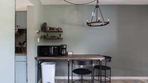 Una cocina compacta con encimera de madera, un microondas negro y una cafetera. Hay dos sillas negras y un cubo de basura blanco.