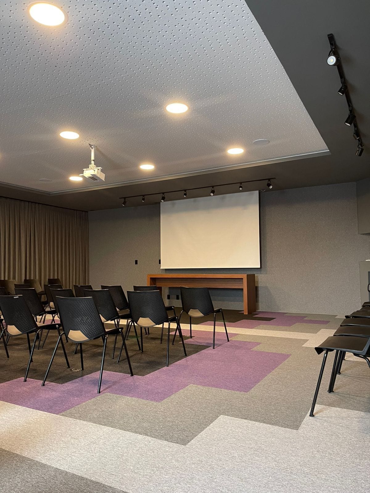 Un salón de conferencias con pantalla de proyector, luces en el techo y filas de sillas.