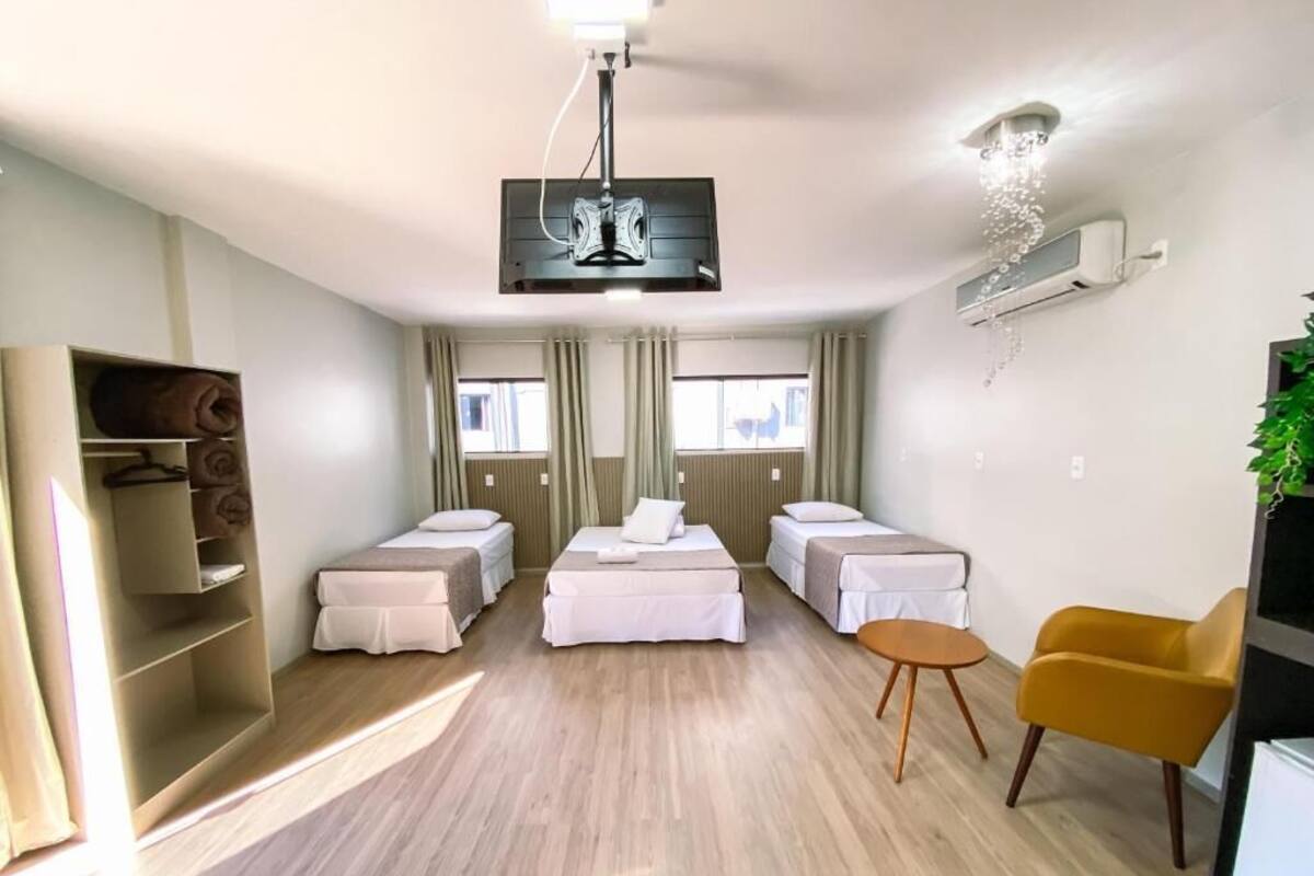 A modern hotel room with a central bed, two smaller beds, a yellow armchair, a TV, and a ceiling fan.