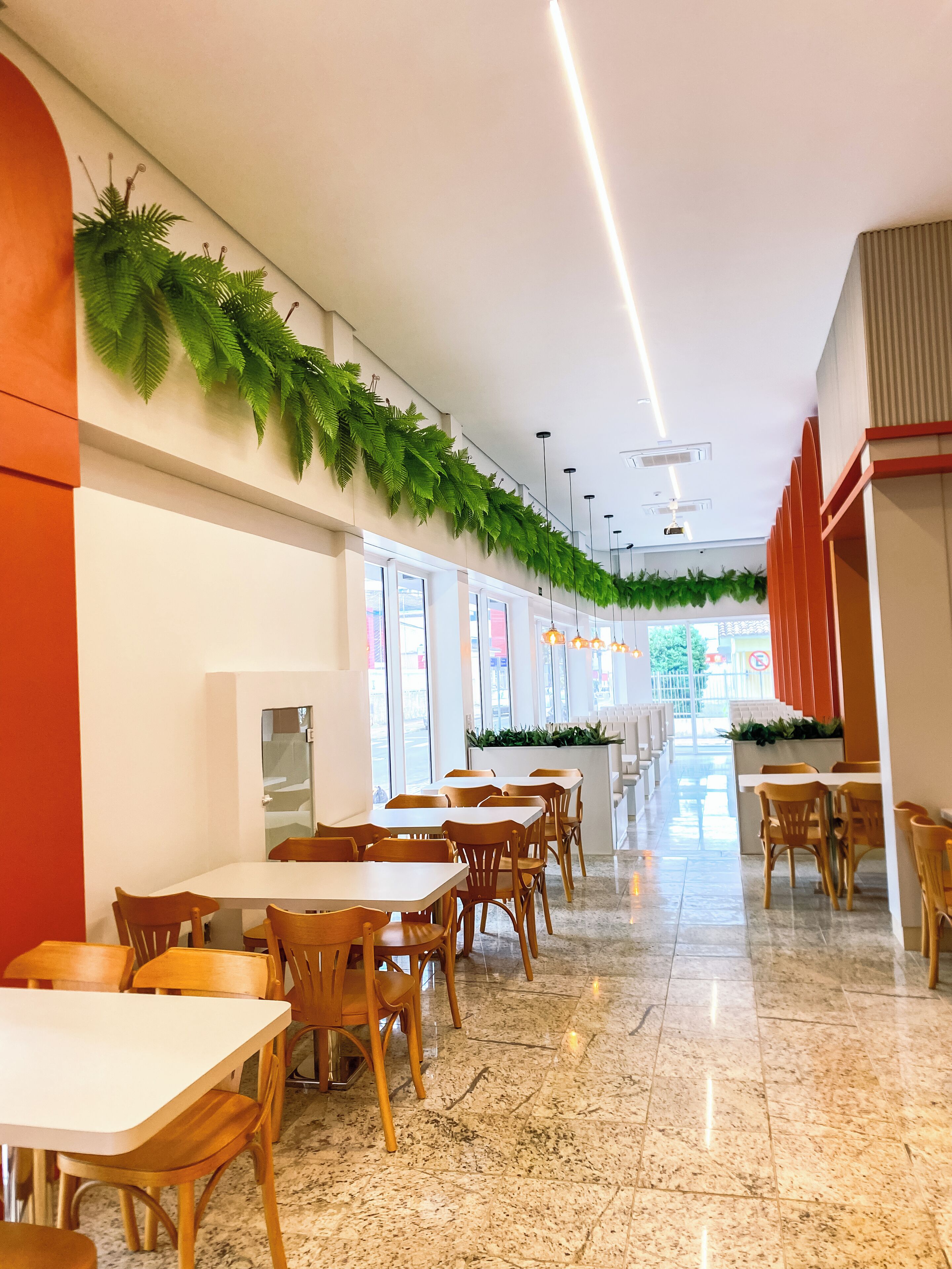 A modern restaurant with long tables and chairs, a tiled floor, and green plants along the walls.