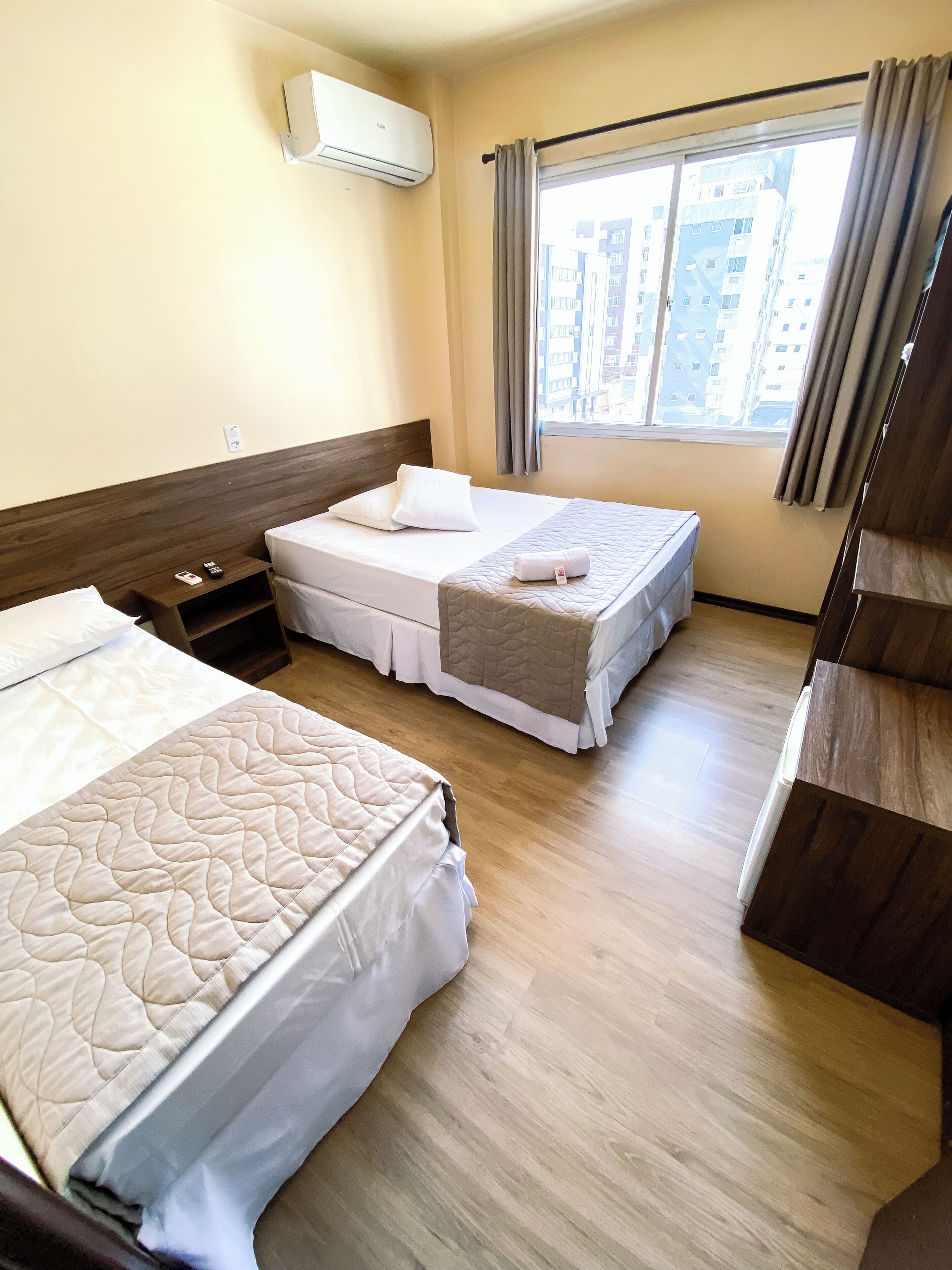 Une chambre d’hôtel avec deux lits, un plancher en bois, une fenêtre avec des rideaux et un climatiseur fixé au mur.