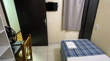 Una habitación pequeña y funcional con cama, una silla, un escritorio y televisor fijado a la pared.