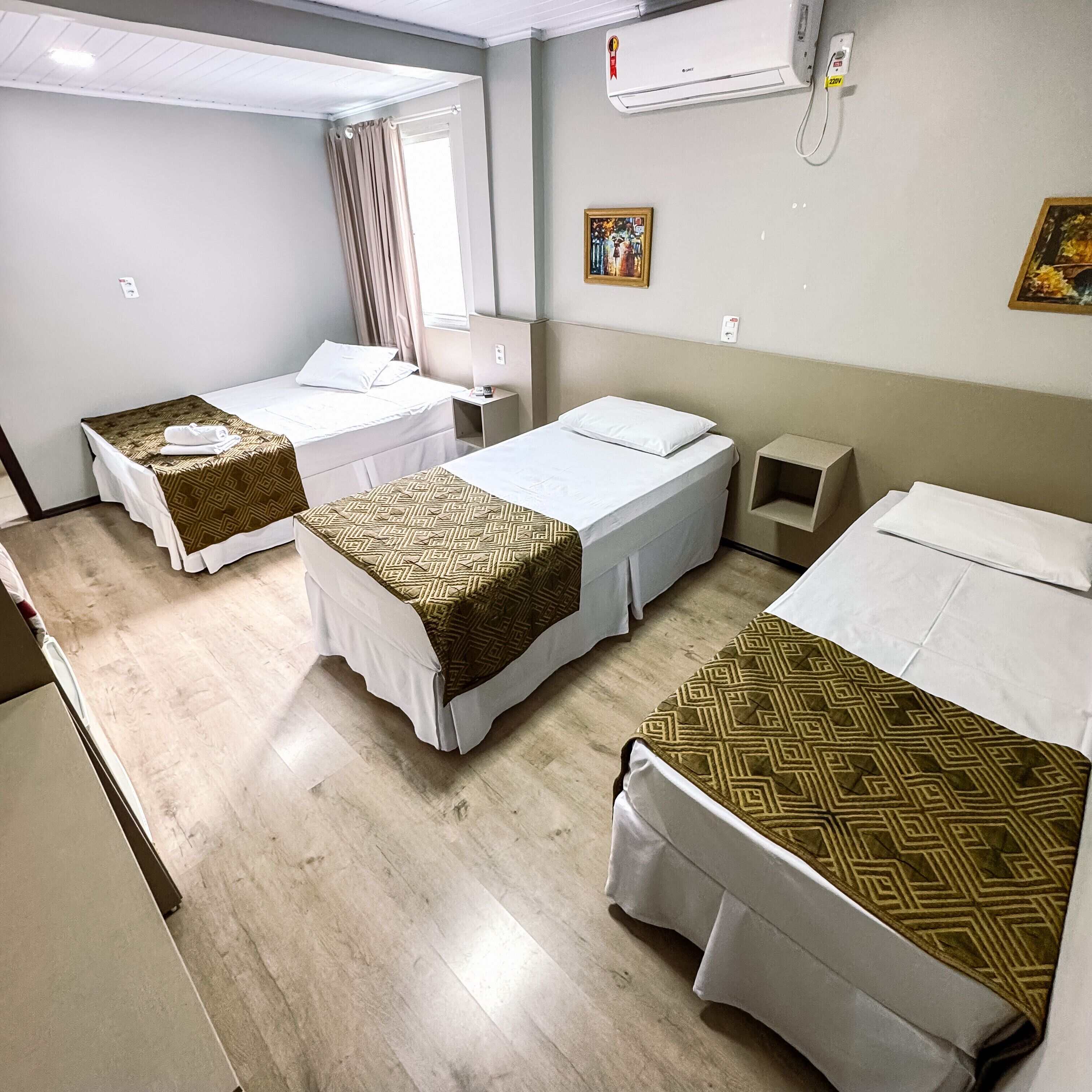 Une chambre d’hôtel avec trois lits simples, une petite table de chevet, une commode, un climatiseur fixé au mur et une fenêtre avec des rideaux.