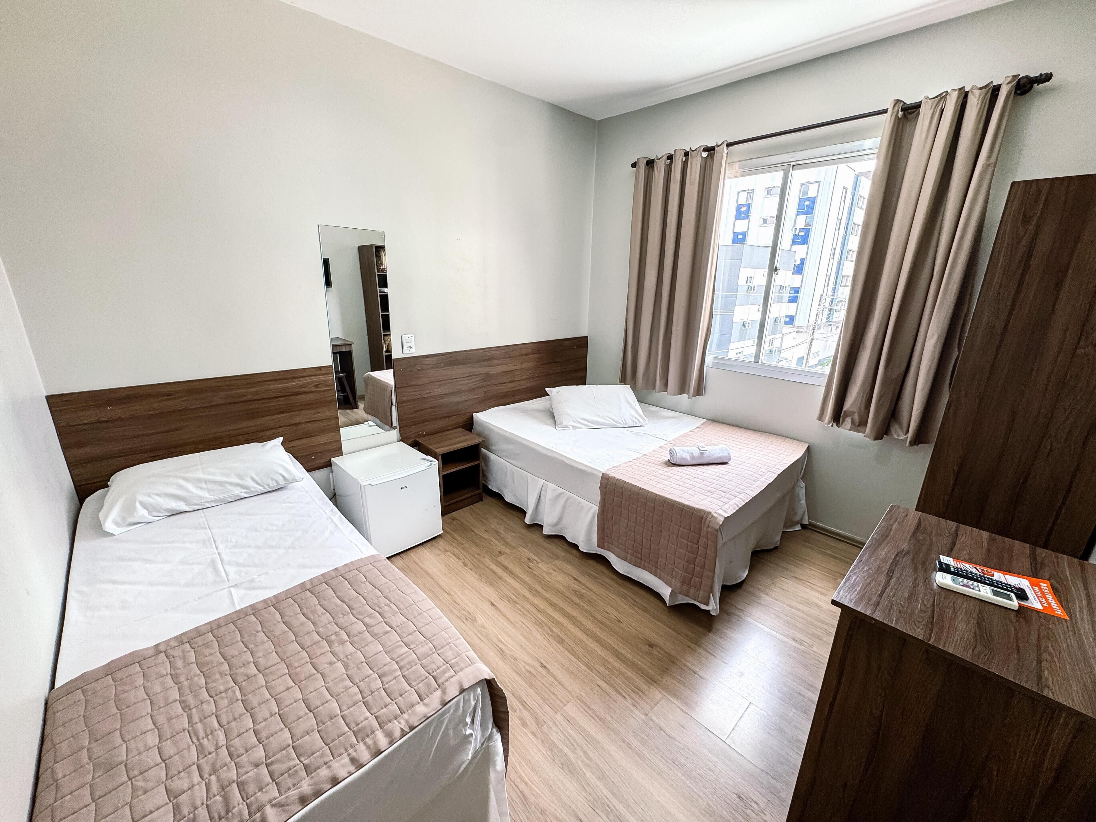 A hotel room with two single beds, a small bedside table, a mirror, a window with curtains, and a wooden desk.