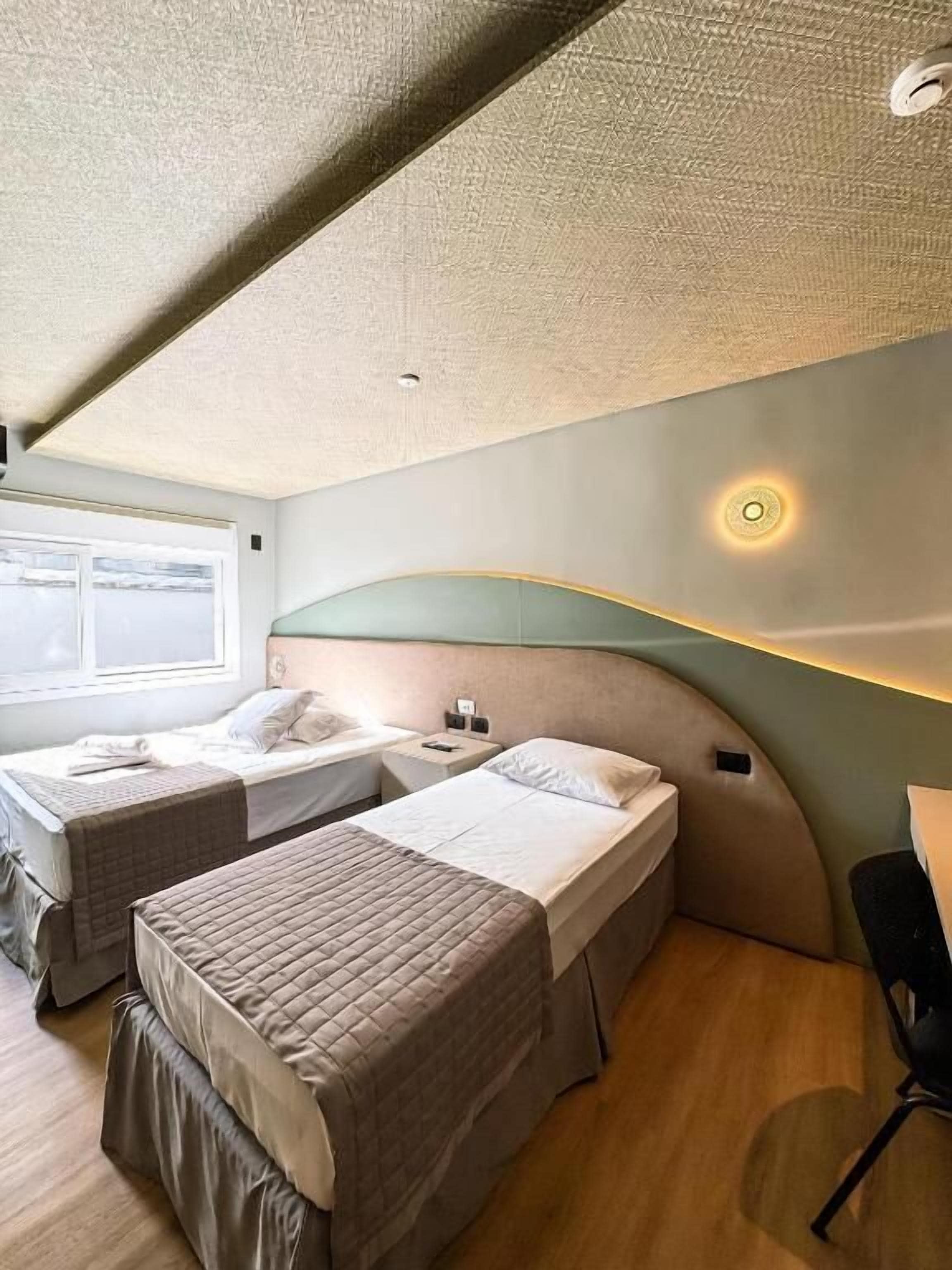 A compact hotel room with two single beds, a small desk, and a window providing a view.
