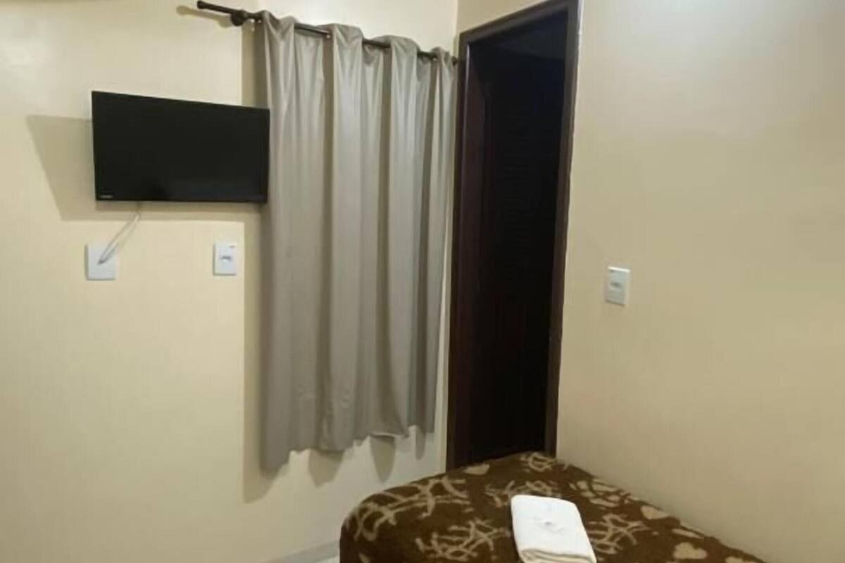 A small, clean room with a bed, a flat-screen TV mounted on the wall, and a window with curtains.