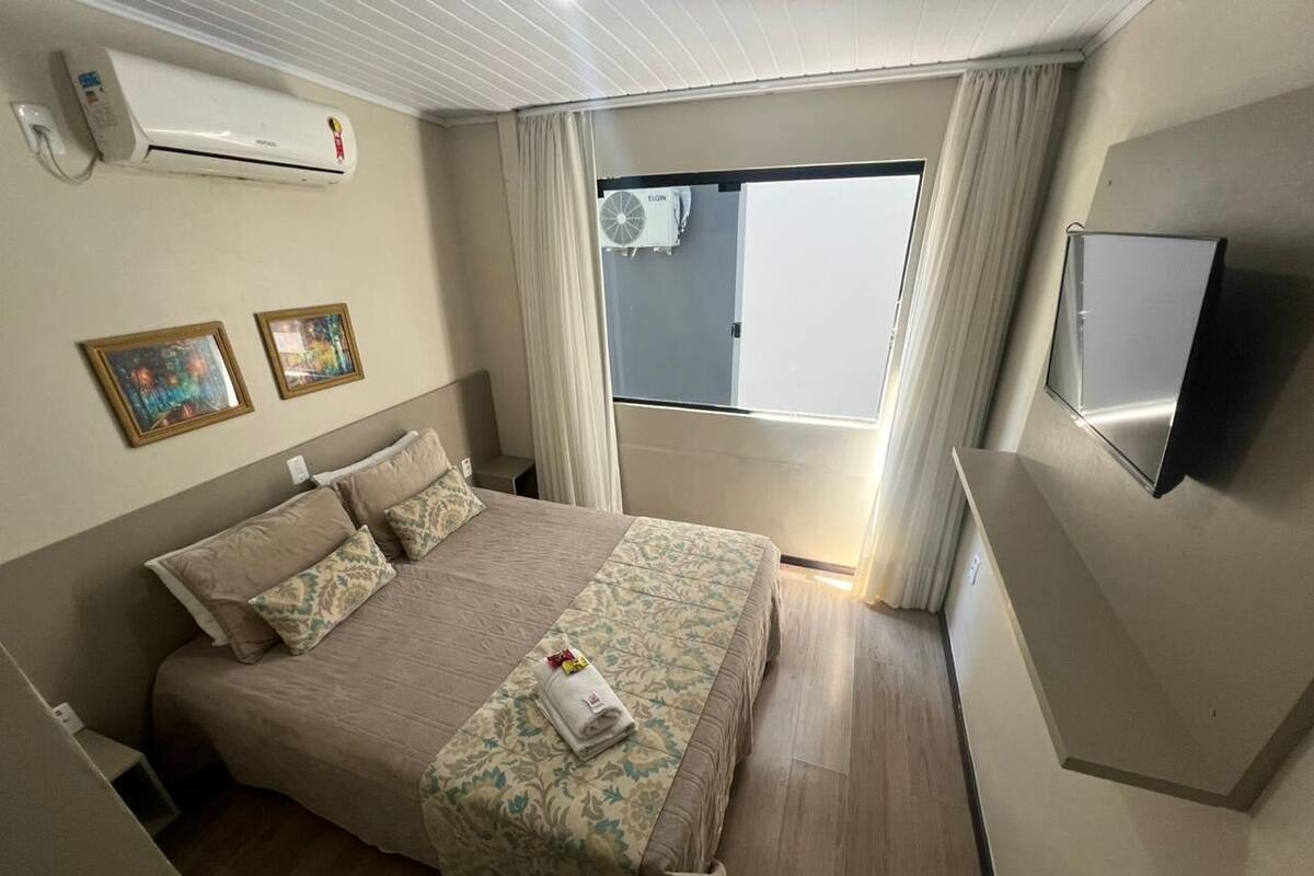 A hotel room with a bed, air conditioning unit, curtains, a television, and a window.