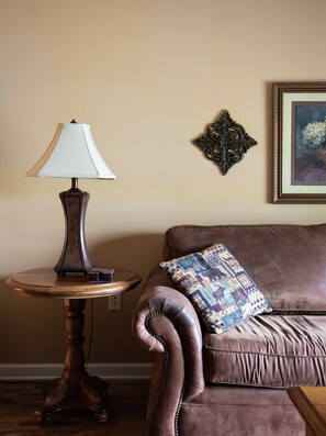 Een kamer met een bruine leren bank, een rond houten bijzettafeltje met een lamp, en een ingelijst schilderij aan de muur.