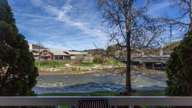 On peut admirer la rivière depuis un balcon doté d’une rambarde en métal, avec une zone herbeuse et un bâtiment au toit rouge en arrière-plan.