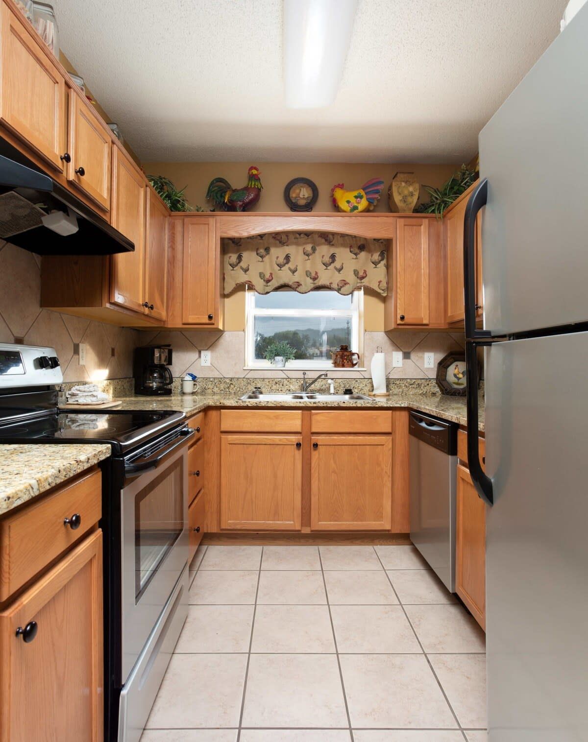 A kitchen with wooden cabinets, a stainless steel refrigerator, a stove, and a window with a rooster-themed curtain.
