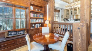 A wooden interior with a round dining table, bookshelves, and a view through large windows.