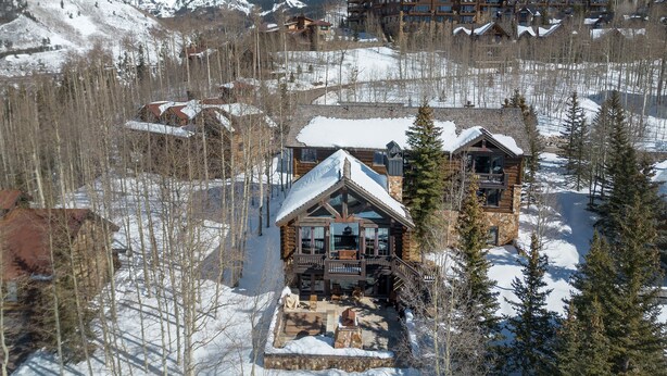 A lodge with a stone facade, large windows, and a balcony overlooking a snowy landscape with mountains in the background.