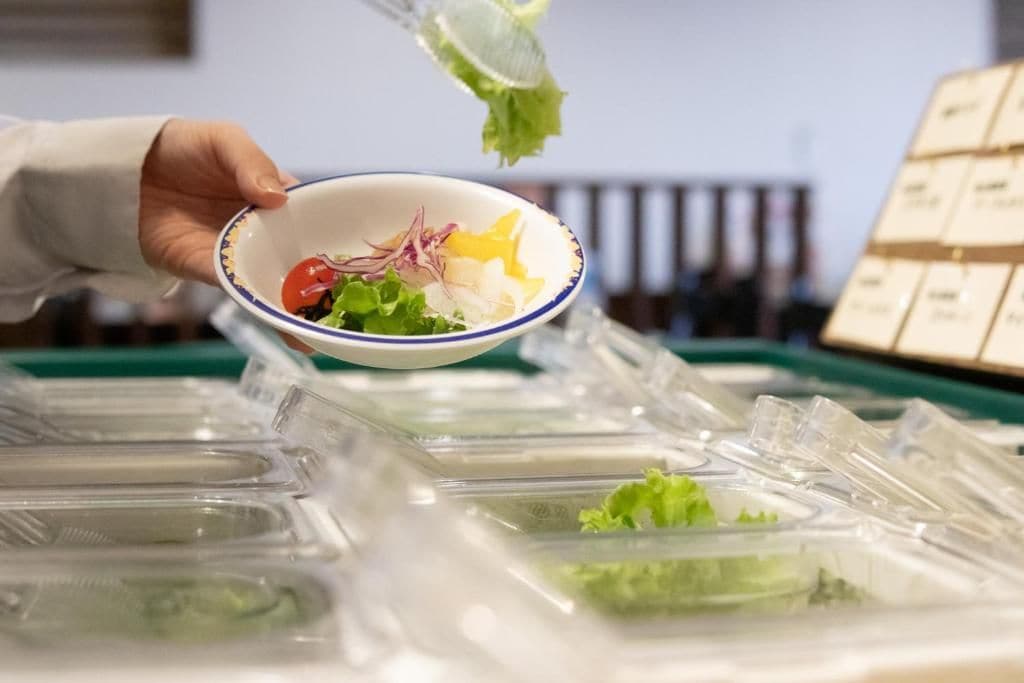 Una mà sosté un bol amb diversos ingredients per a amanides, damunt d'una safata amb recipients de plàstic transparent.
