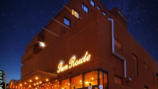 A building with a neon sign reading "Sun Route" at night, featuring a staircase leading to the entrance and a parking area visible below.