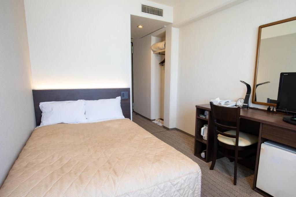 A hotel room with a bed, a desk with a chair, a mirror, and a small refrigerator.