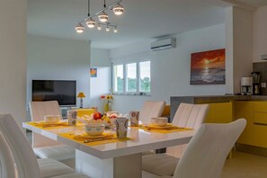 Uma sala de jantar moderna com uma mesa branca, jogos americanos amarelos e cadeiras brancas. Há uma televisão ao fundo, um quadro na parede e uma área de cozinha com armários amarelos.