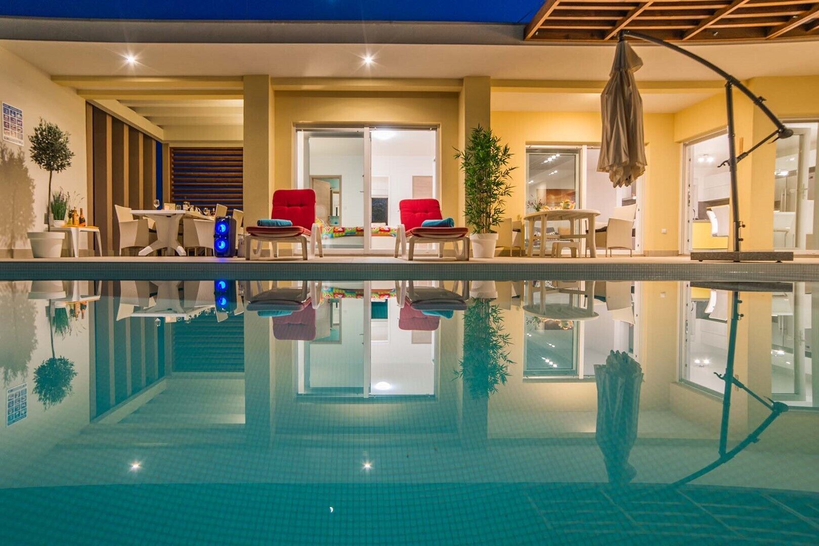 A swimming pool with lounge chairs and tables, surrounded by a modern interior with a ceiling fan and potted plants.