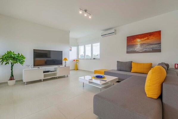 A modern living room with a flat-screen TV, a sofa with yellow cushions, a coffee table, and a potted plant.