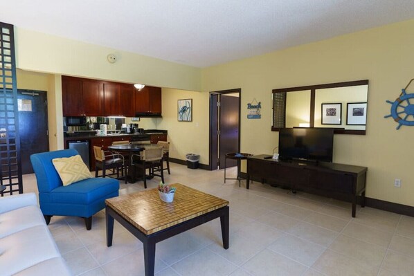 A living room with a blue sofa, a coffee table, a TV, and a kitchen area with wooden cabinets.
