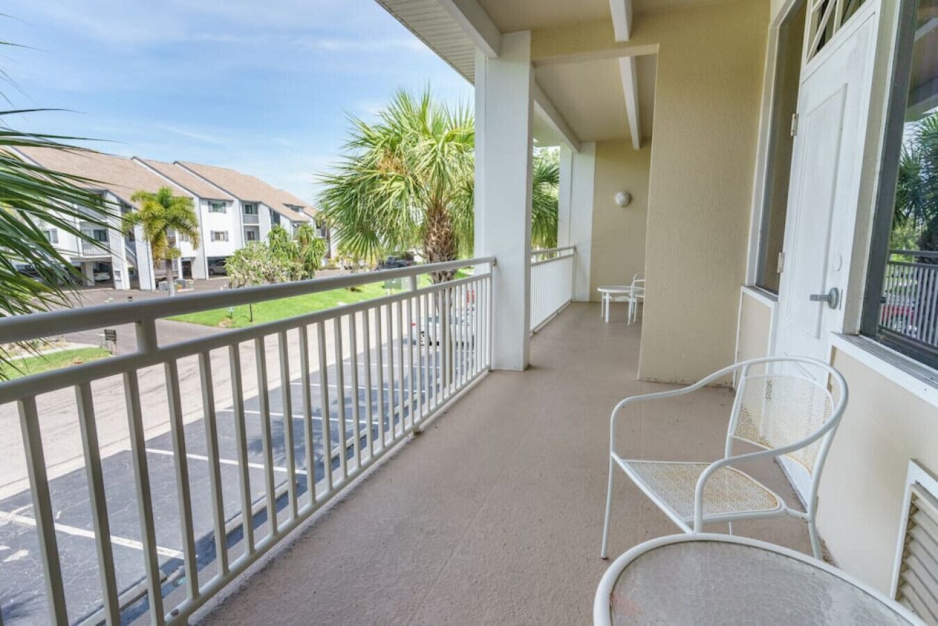 A balcony with a white railing, a white chair, and a view of a street with palm trees and buildings.
