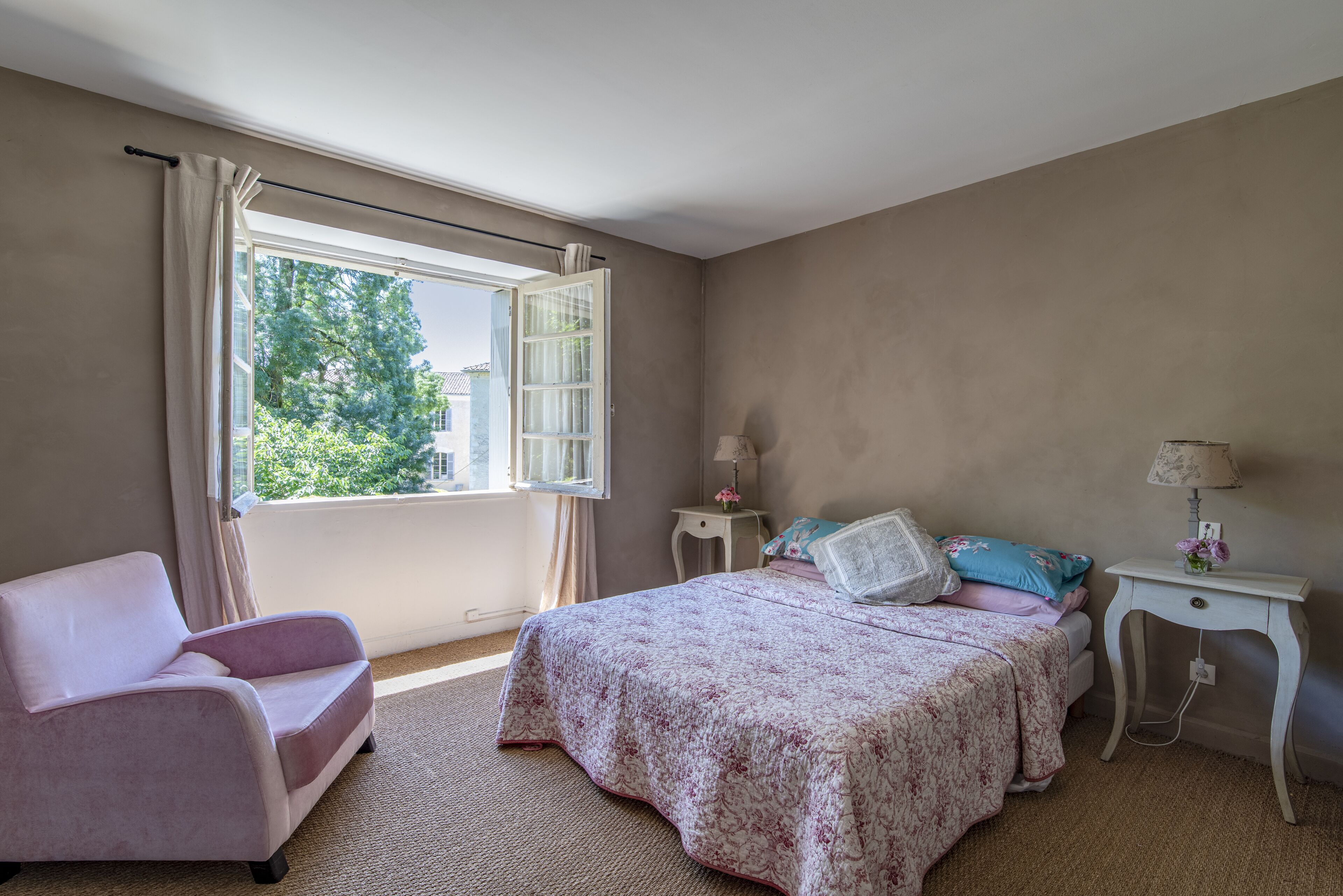 A bedroom with a bed, a chair, a window with curtains, and a nightstand with a lamp.