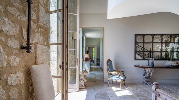 Une pièce aux murs de pierre, avec une grande fenêtre, une chaise, une table et un escalier menant à une autre pièce.