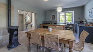 Un coin repas avec une grande table en bois et des chaises en osier, un poêle noir et des luminaires suspendus.