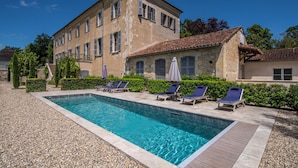 Une piscine entourée de chaises longues et de parasols, elle-même bordée d’un bâtiment en pierre doté de nombreuses fenêtres et d’un toit recouvert de tuiles.