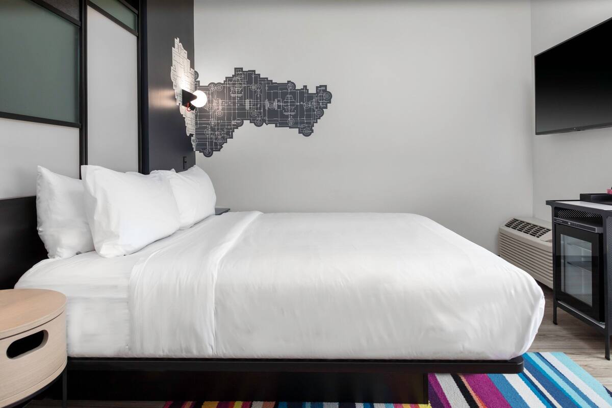 A modern hotel room with a large bed, a wall-mounted TV, and a unique cityscape artwork.