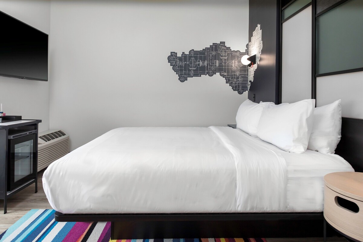 A modern hotel room with a large bed, a flat-screen TV, a wall-mounted light fixture, and a small desk with a computer.