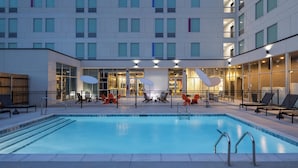 Zona de piscina hotelera con una alberca rectangular, camastros y un edificio con múltiples ventanas y balcones.