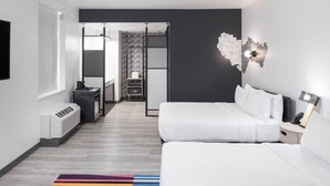Habitación de hotel moderna con cama blanca, escritorio, silla, calefactor, mesita de noche y lámpara de pared.