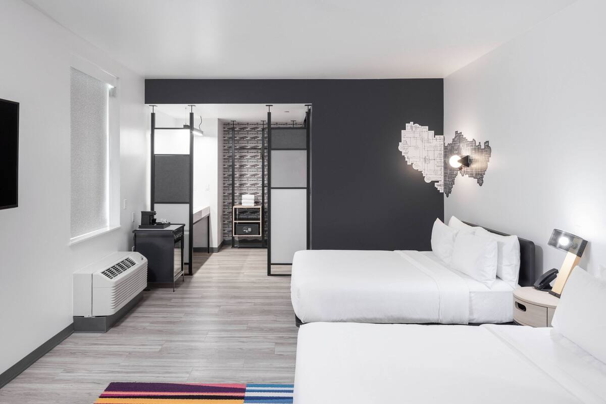 Une chambre d’hôtel moderne avec un lit blanc, un bureau, une chaise, un radiateur, une table de chevet et une lampe fixée au mur.