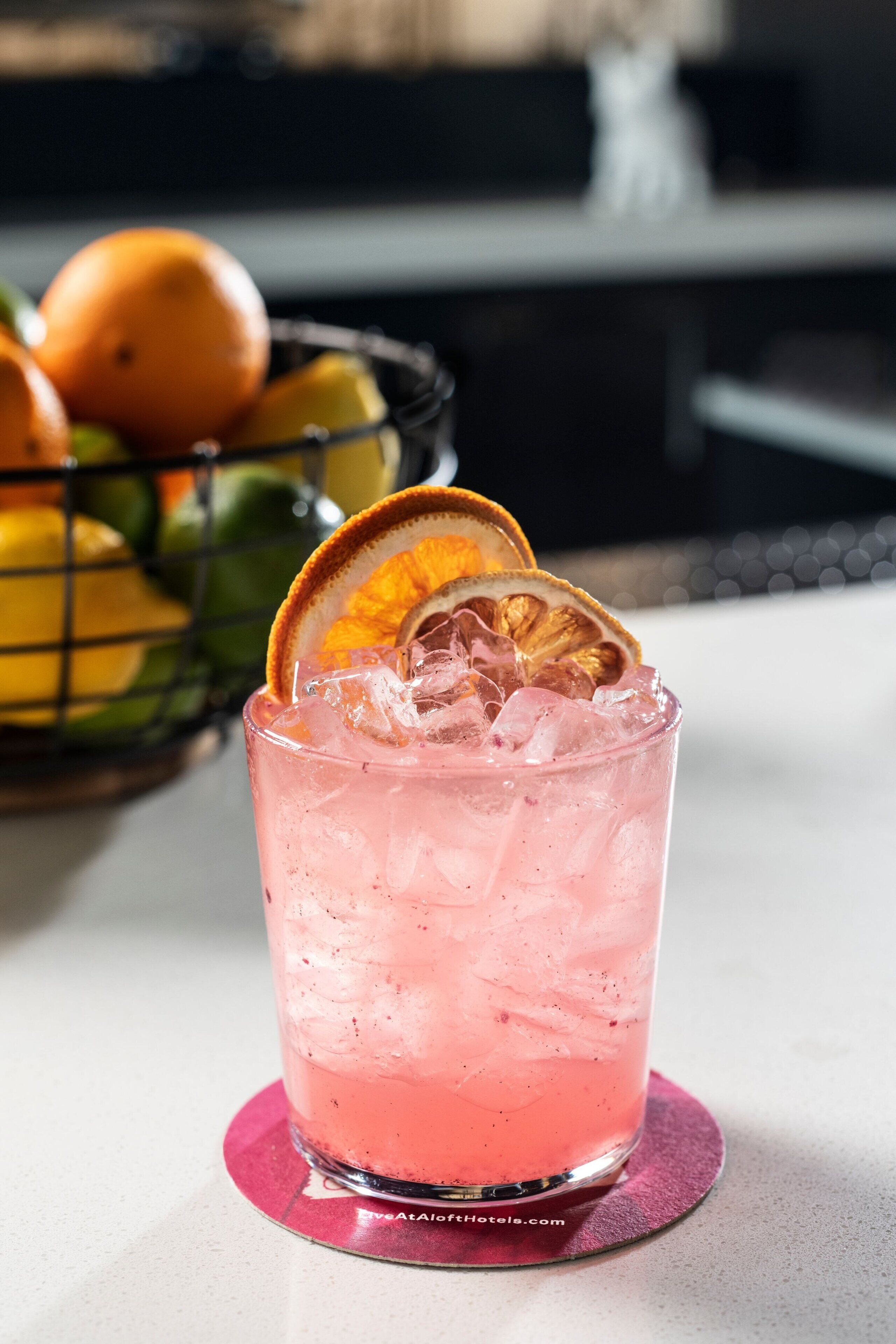 A pink cocktail with ice and a slice of orange in a glass on a coaster.