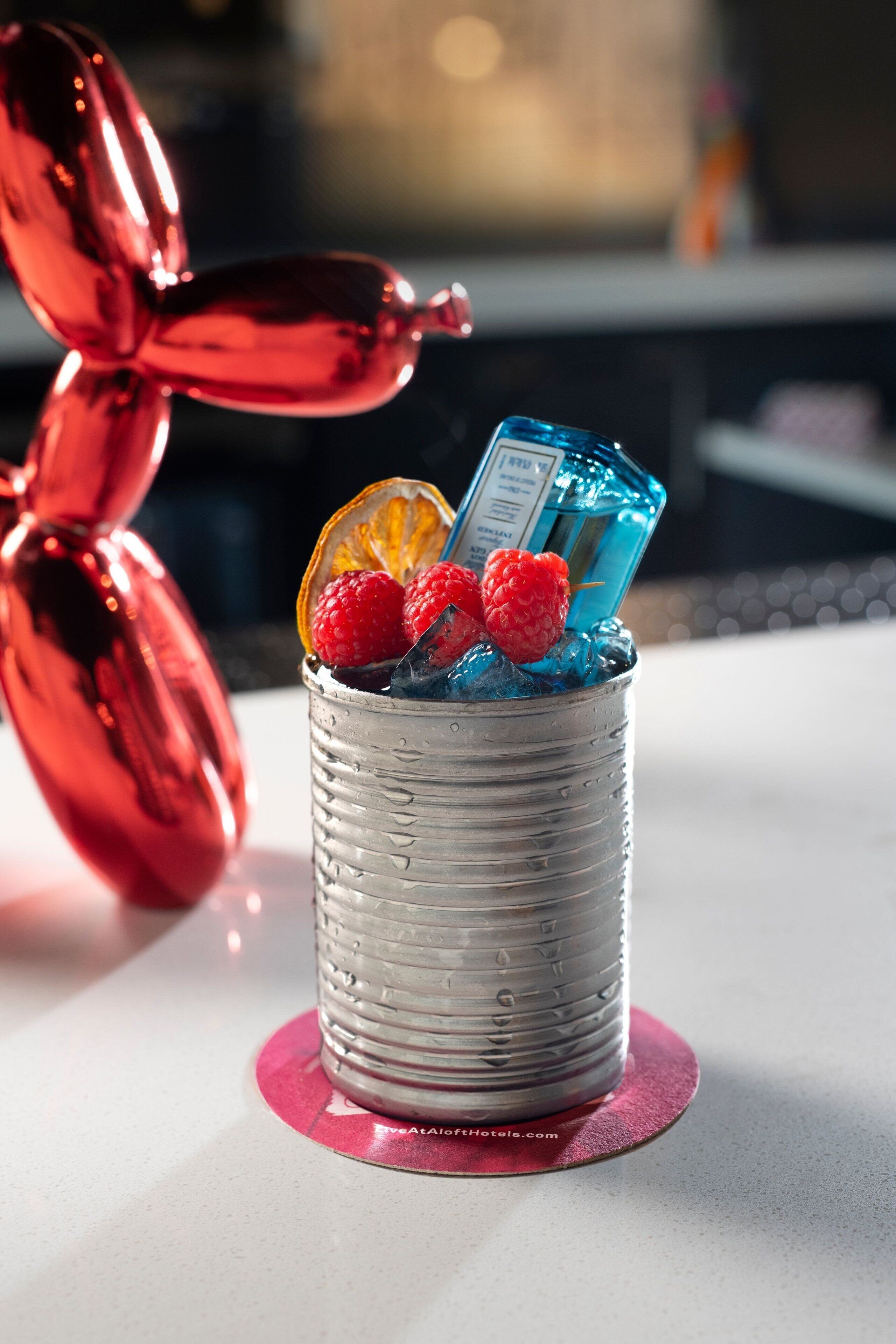A cocktail in a metal cup with a red balloon and a blue bottle in the background.