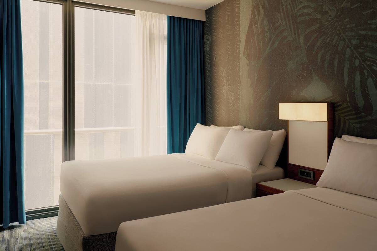 Een hotelkamer met twee bedden, een groot raam met gordijnen, een wand met bladmotief en een bedlamp.